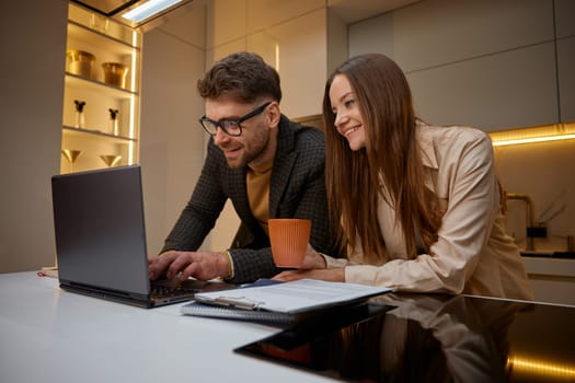 Young family using laptop computer on home kitchen. Husband helping his loving wife with online project showing life hack operating with data and software