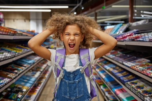 Stressed girl child feeling crazy during busy shopping at stationery shop. Mad female kid tearing hair on head screaming loudly portrait. Children emotion, purchases and back to school concept