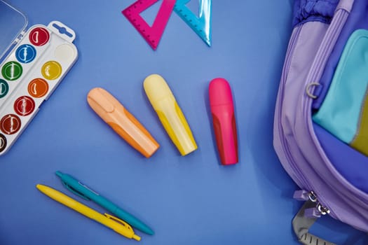 Backpack with watercolor paints, markers and ballpoint pens colorful school supplies on blue background. Back to school flat lay copy space