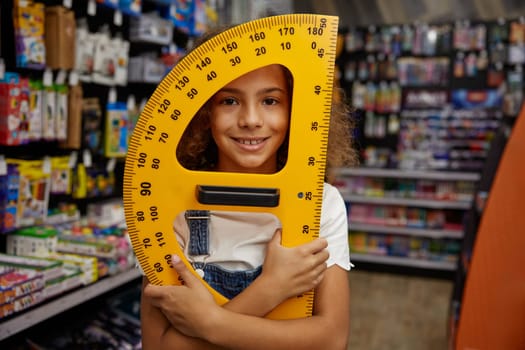 School girl student holding huge protractor ruler stationery in hands while standing at store. Creative studying tools for education