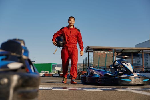 Equipped go-kart driver wearing overalls and holding helmet walking to car before race at starting line on motor racing track. Automotive sports activity, carting championship