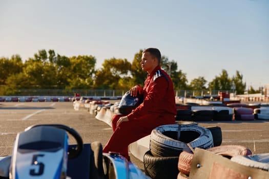 Back view shot of male racer in protective overalls holding helmet taking rest while training at go-kart track outdoors