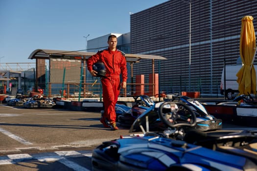 Well-equipped go-kart driver wearing overalls and holding helmet walking to car before race at starting line on motor racing track. Automotive sports activity, carting championship