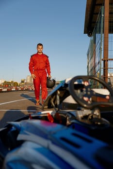 Confident go-kart driver wearing overalls and holding helmet walking to car before race at starting line on motor racing track. Automotive sports activity, carting championship