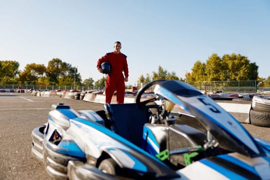 Well-equipped go-kart driver wearing overalls and holding helmet walking to car before race at starting line on motor racing track. Automotive sports activity, carting championship