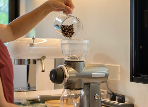 Closeup of female hand pouring out coffee beans into coffee grinder machine in the kitchen at home