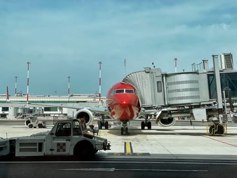 Passenger airplane with a red nose cone docked at the airport terminal gate with jet bridge attached, ready for boarding