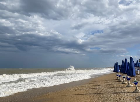 Dramatic cloudy sky over rough sea waves, with a line of closed blue umbrellas along a deserted beach  concept of off-season travel