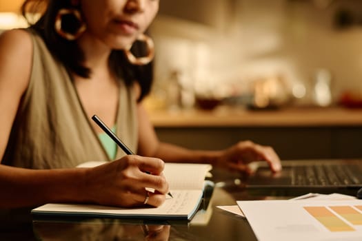 Hand of young modern businesswoman with pen making notes on page of copybook while sitting in front of camera by workplace with papers
