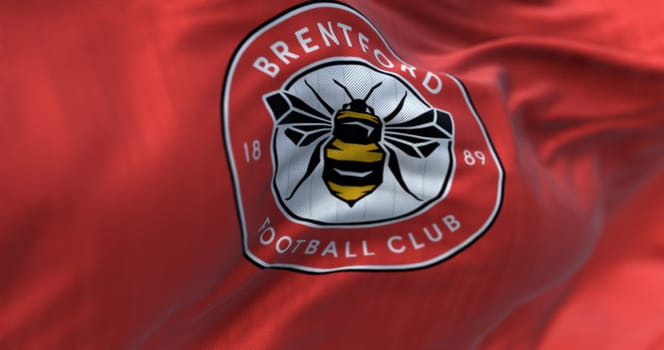 London, UK, Feb 19 2023: Close-up of Brentford Football Club flag waving. Circular emblem with a bee inside on red. Illustrative editorial 3d illustration render. Textured background. Selective focus