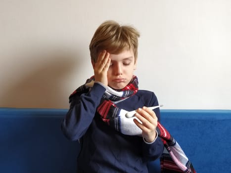 The boy got sick, his head hurts. Viral disease is progressing. The childs neck is wrapped in a warm scarf. The kid looks dejectedly at the thermometer, holds his hand on his forehead and suffers.