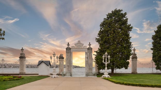 Ornate gates of Dolmabahce Palace open up to a breathtaking sunset over waters of Bosphorus Strait in Istanbul, Turkey, flanked by trees and intricately designed fence posts against the dramatic sky