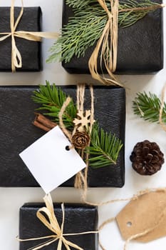 Black wrapped present with Christmas square gift tag with fir tree branches, pine cones and holiday decorations, top view. Rustic winter composition, Mockup, copy space