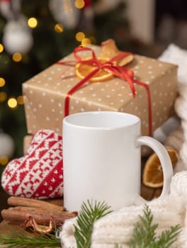 White coffee mug near Christmas present, cosy heart and sweater close up against Christmas tree lights and burning fireplace, copy space. Warm winter family atmospheric mock up, magical Holiday time