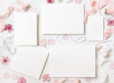 Paper cards near pink decor, flowers, seals and silk ribbons on white table top view, wedding suite mockup. Romantic scene with blank cards flat lay. Valentines, Spring or Mothers day concept