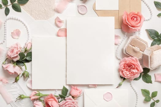 Blank cards near light pink roses, engagement ring in a box and silk ribbons on white table top view, wedding mockup. Romantic flat lay with vertical cards and pastel decor