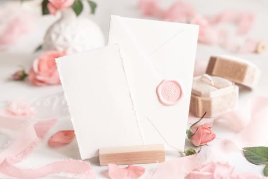 Card and sealed envelope near light pink roses, petals, engagement ring in a box and silk ribbons on white table close up, wedding mockup. Romantic scene with pastel decor