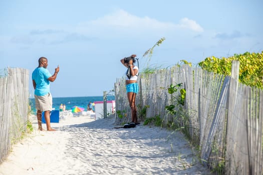 Miami, USA - August 23, 2014: couple at south beach passing the protection dune in late afternoon. He uses his mobile, she prepares her hair.