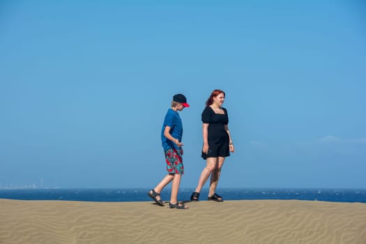 Two teenagers stand on a sand dune, with sea and blue sky