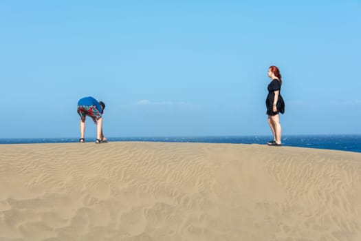 Two teenagers stand on a sand dune, with sea and blue sky