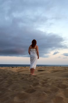 A young girl stands in a white dress in the sand dunes with sky and clouds in the late afternoon