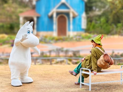 japan, saitama - mar 26 2023: Moomintroll costume waving hand to his smiling friend Snufkin wearing a green hat carrying a backpack with a camp mattress smoking a pipe on a bench in Moominvalley park.