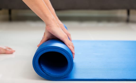 Cropped image of woman rolling blue mat after doing yoga exercise workout in living room at home. Healthy life and fitness training workout concept
