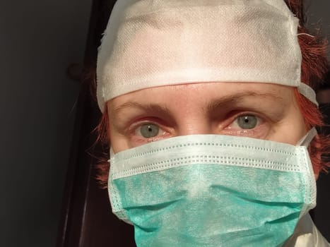 Redhead woman with sick tired eyes in a protective surgical mask of green color. Gray eyes and red hair. Dark background. Protective measures against viral and bacterial diseases