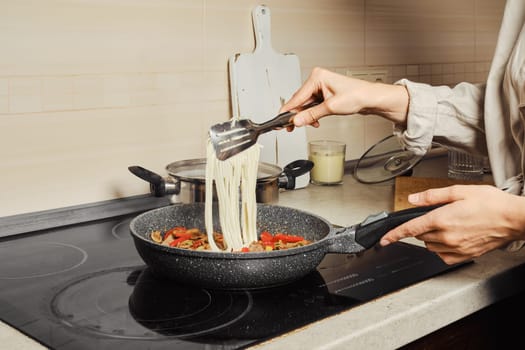 Unrecognizable woman transfers noodles with kitchen tongs into a frying pan with vegetable dressing and beef