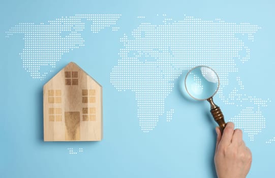 Wooden house on a world map background with a person's hand holding a magnifying glass, symbolizing the concept of searching for a country to move to