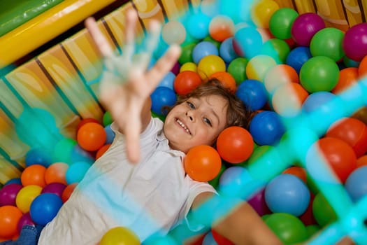 Happy boy child playing in basin filled with multi colored plastic ball. Excited cheerful kid having fun in big dry paddling pool at indoor playroom