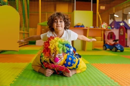 Portrait of happy smiling clown and little boy child lying on floor of indoor playground. Show entertainment, animation program for kids to celebrate birthday party with fun and joy