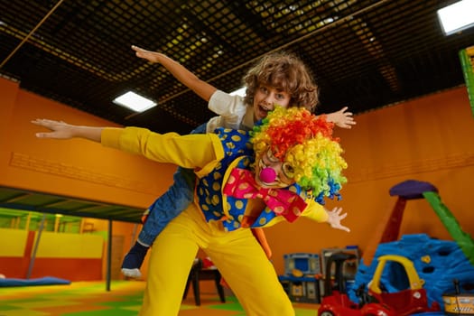 Happy children playing with male clown wearing funny colorful costume standing over indoor playroom interior. Positive preschooler boy and animator laughter enjoying games and joyful reaction time