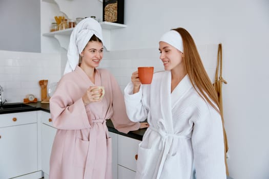 Happy girls wearing bathrobe with wrapped towel on head standing and chatting on home kitchen. Student hostel space sharing and friendship concept