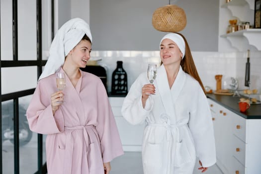 Two young woman in bathrobe with wrapped towel on head standing in living room clinking wineglasses celebrating pajama party. Free time leisure activity at student hostel