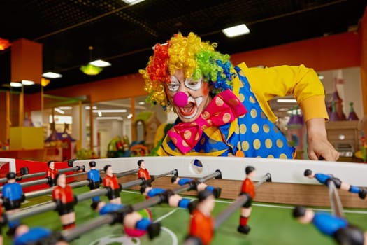 Happy excited clown with funny makeup playing table football in entertainment center indoors. Creative people positive emotion and leisure activity concept