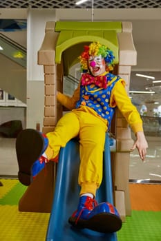 Overjoyed clown in colorful costume sliding down at child playroom. Creative animator feeling fun, playing at kids entertainment center performing games at birthday party