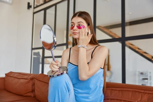 Young woman applying eye patch looking at mirror while sitting on sofa at cozy living room. Home beauty procedure