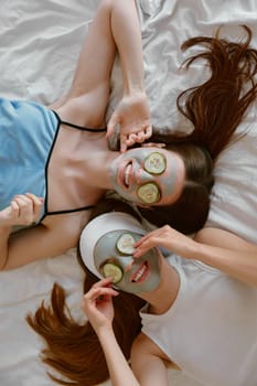 Overhead view on funny young girl friends applying clay mask and cucumber eye patch for beauty wellness during spa day at home