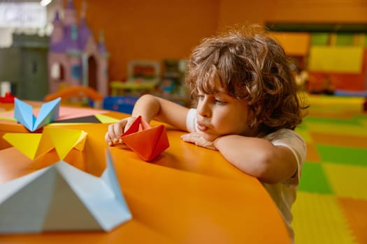 Cute boy child playing handmade paper boat at childcare center playroom. Enthusiastic male kid constructing origami toys enjoying leisure activity at amusement area