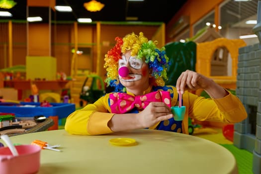 Funny clown wearing multi-colored wig and costume playing with toy tea set at kids playroom. Birthday party animation show concept