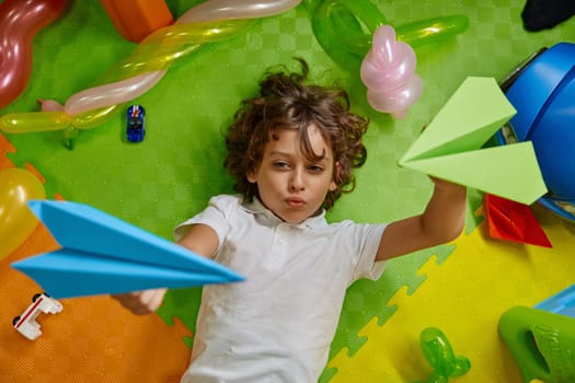 High angle view on little boy kid playing paper planes on cozy foamy floor of childcare center. Playful time for children at kindergarten or playroom in shopping mall