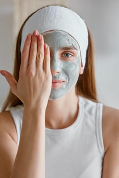Portrait of young woman applying clay facial cosmetology mask covering half face with hand. Healthy skin and beauty, home spa procedure with natural cosmetics concept