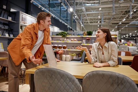 Positive loving couple testing new furniture for dining room at home furnishing store department. Cheerful man and woman buyers having fun during shopping