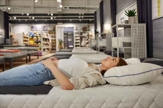 Young woman shopper choosing new bed at modern furniture showroom. Attractive female customer testing comfortable bedstead enjoying shopping activity on weekend