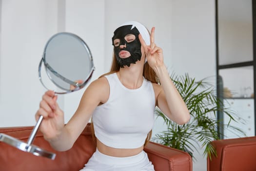 Young woman applying facial mask having fun grimacing looking at mirror. Home beauty skin care procedure concept