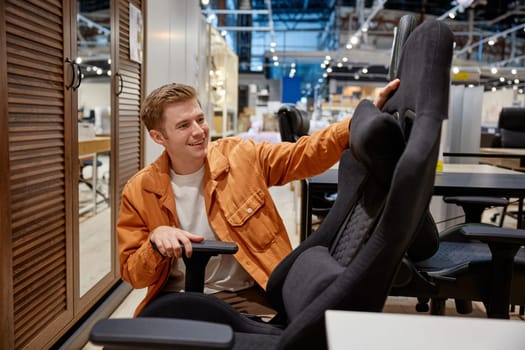Young handsome man making decision what home office armchair to buy at shop store. Low price versus comfort and high quality