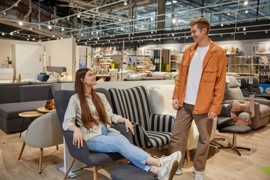 Young family couple choosing comfortable chair for home interior at shop store with great assortment and bog discounts. Attractive woman testing armchair in furniture retail market