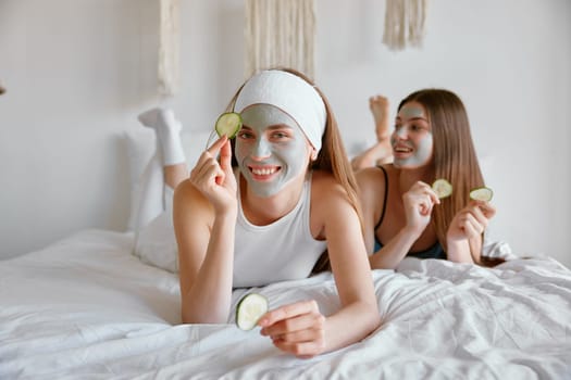 Portrait of two girl friends celebrating home spa party together applying cucumber slices as eye patches and cosmetic clay facial mask. Beauty and skincare concept