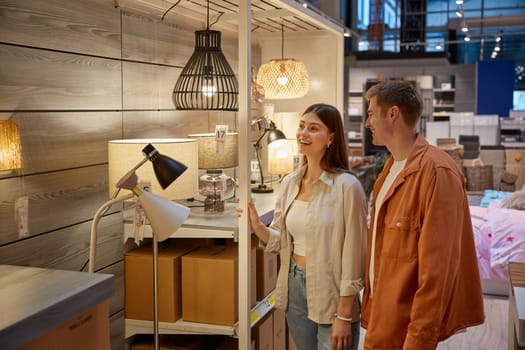 Young family couple choosing chandelier in building shop center. Handsome man and attractive woman lovers selecting exclusive electric lightning equipment for their new apartment house at retail store